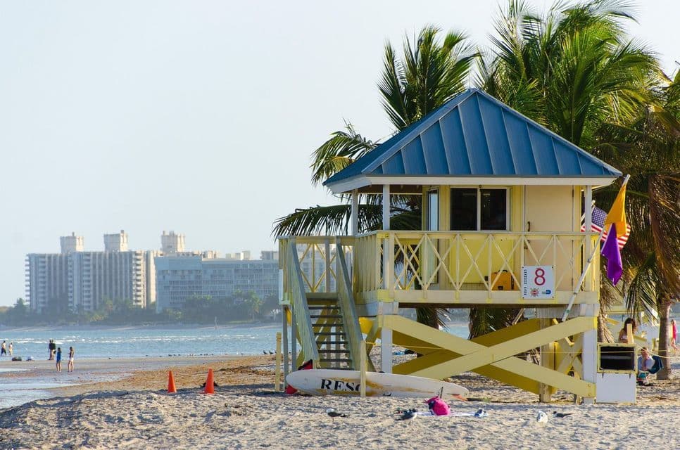 Una torre di salvataggio gialla con tetto blu si trova su una spiaggia sabbiosa, con palme e uno skyline della città sullo sfondo.