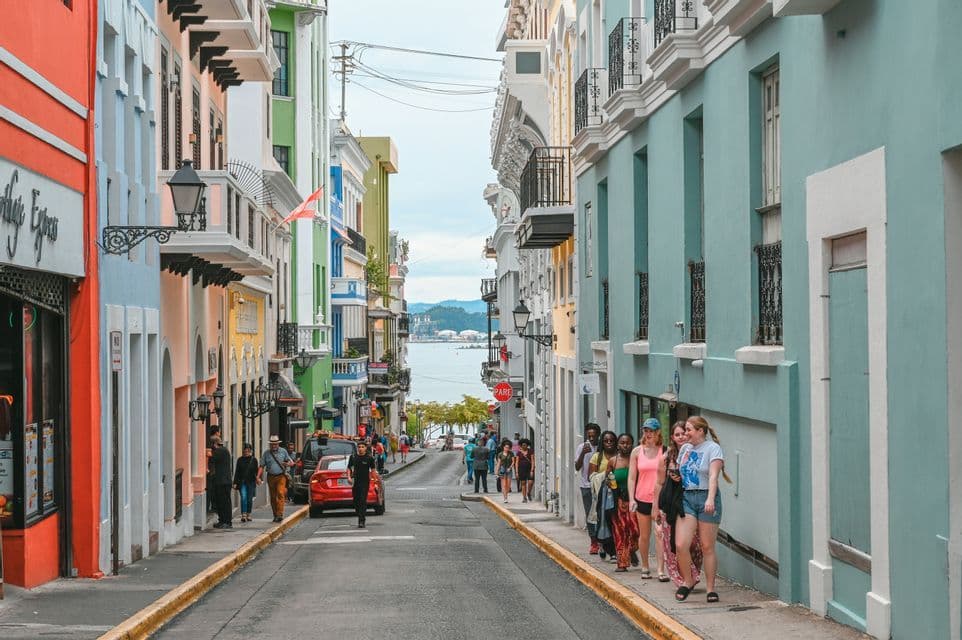 Un gruppo WeRoad passeggia lungo il marciapiede di una strada in pendenza, fiancheggiata da edifici colorati, con il mare visibile sullo sfondo.