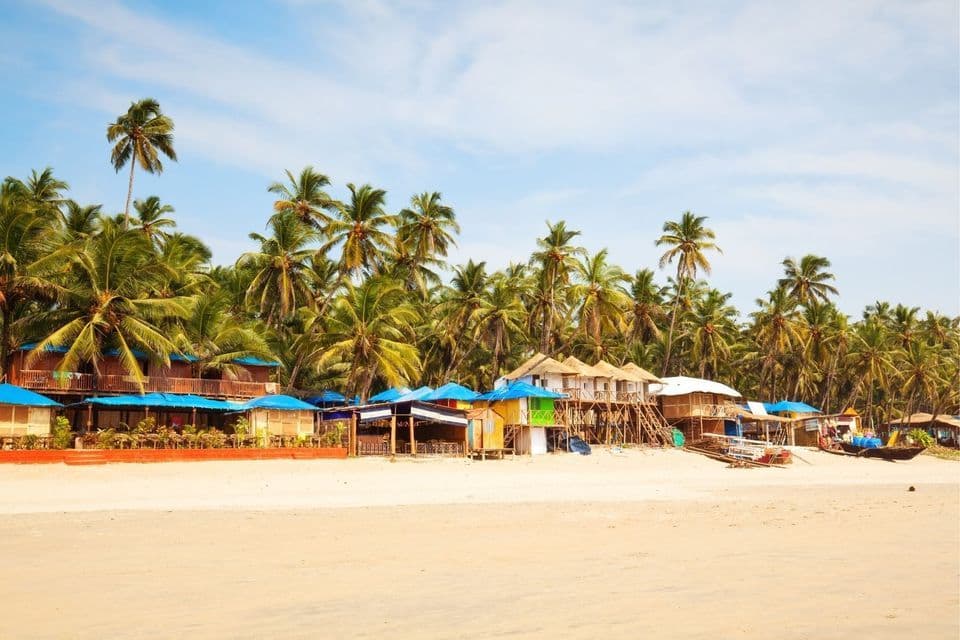 Bunte Strandhütten und Buden säumen einen breiten Sandstrand, dahinter ein dichter Palmenhain.