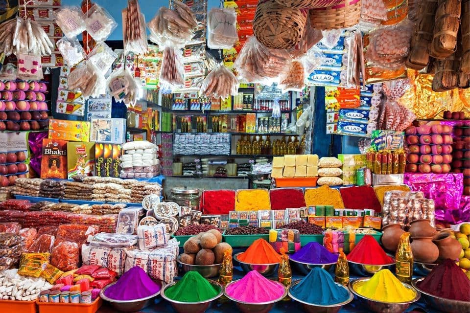 Ein hell beleuchteter Marktstand ist gefüllt mit bunten Pulvern in Schalen, Gewürzen, Obst und anderen Waren zum Verkauf.