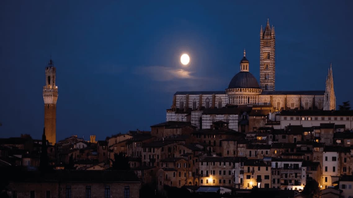 Una città storica con una cattedrale e torri illuminate, sotto un cielo notturno buio con una luna piena splendente.