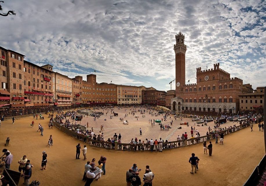 Una vista panoramica di una grande piazza pubblica non lastricata, gremita di persone, circondata da edifici storici e da un'imponente torre dell'orologio sotto un cielo nuvoloso.