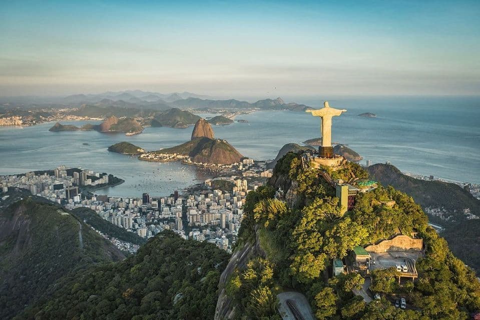 Une grande statue du Christ aux bras ouverts se dresse sur une montagne verdoyante, surplombant une ville côtière et une baie au lever du soleil.