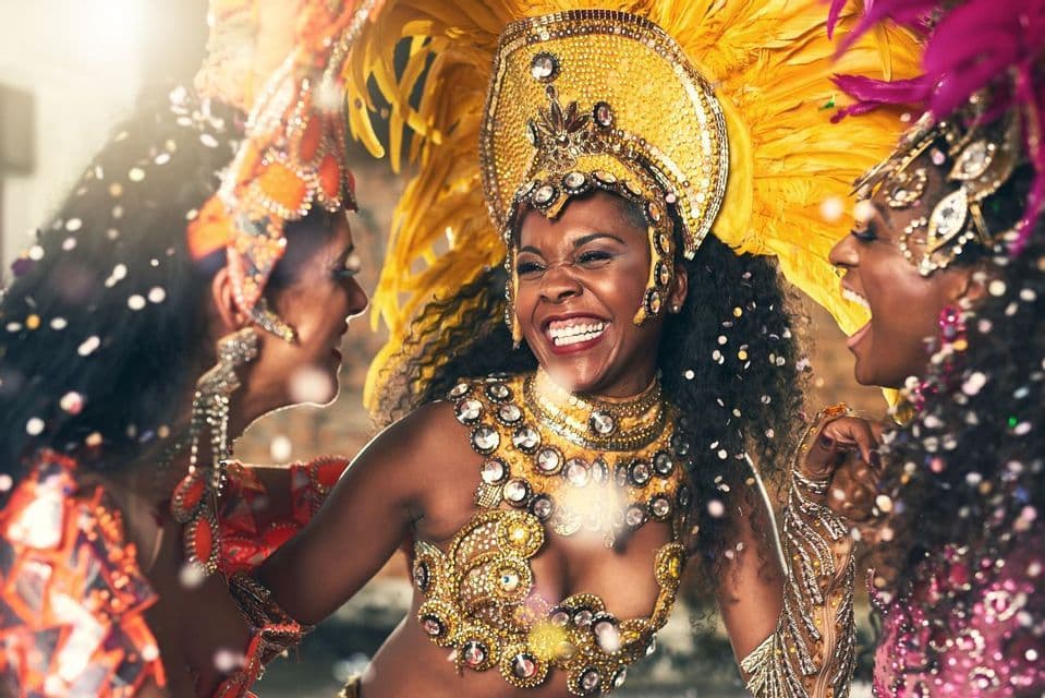 Trois femmes rient et dansent ensemble, vêtues de somptueux costumes de carnaval à plumes, au milieu de paillettes qui tombent.