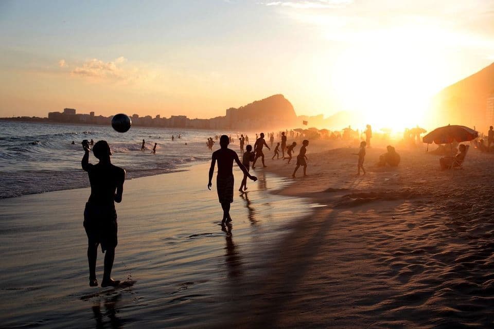 Silhouettes de personnes jouant au ballon sur une plage bondée au coucher du soleil, avec une ligne d'horizon urbaine en arrière-plan.