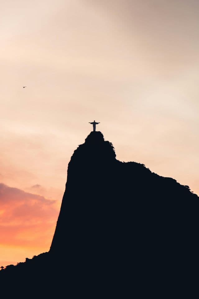 Silhouette d'une grande statue aux bras tendus sur un sommet de montagne, devant un ciel de coucher de soleil aux tons chauds.