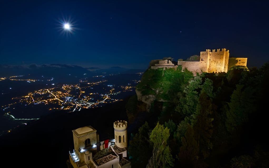 Un castello illuminato si erge in cima a una scogliera, affacciato su una valle piena di luci cittadine, sotto una luna splendente di notte.