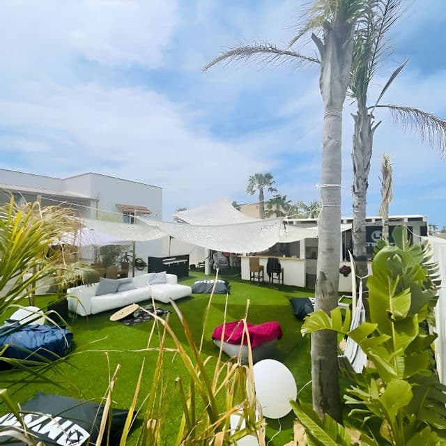 Un'area lounge esterna con erba sintetica, un divano bianco, pouf e vele ombreggianti bianche, sotto palme e un cielo parzialmente nuvoloso.