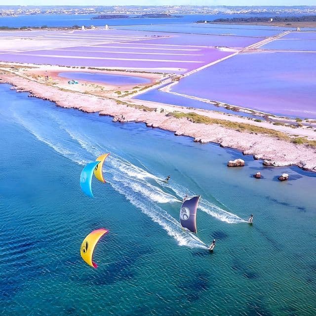 Una vista aerea di diversi kitesurfer su acqua blu accanto a una costa con vaste saline rosa e viola.