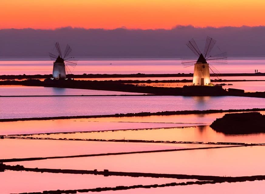 Due mulini a vento illuminati si ergono tra le saline al tramonto, riflettendosi nell'acqua sotto un cielo arancione e viola.