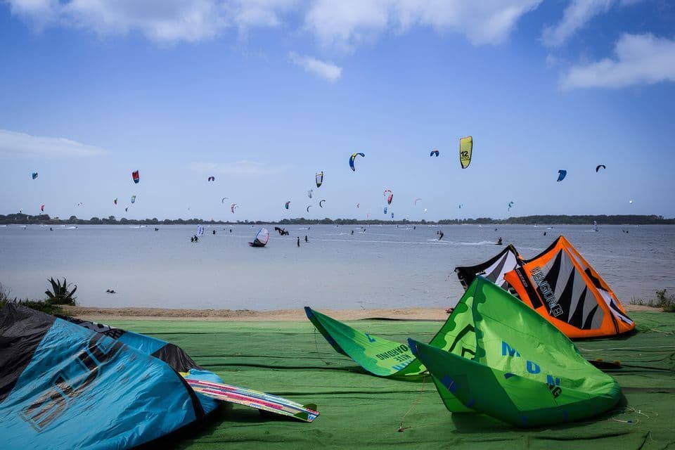 Attrezzatura da kitesurf colorata poggia su un telone verde vicino all'acqua, mentre numerose persone fanno kitesurf sullo sfondo sotto un cielo nuvoloso.