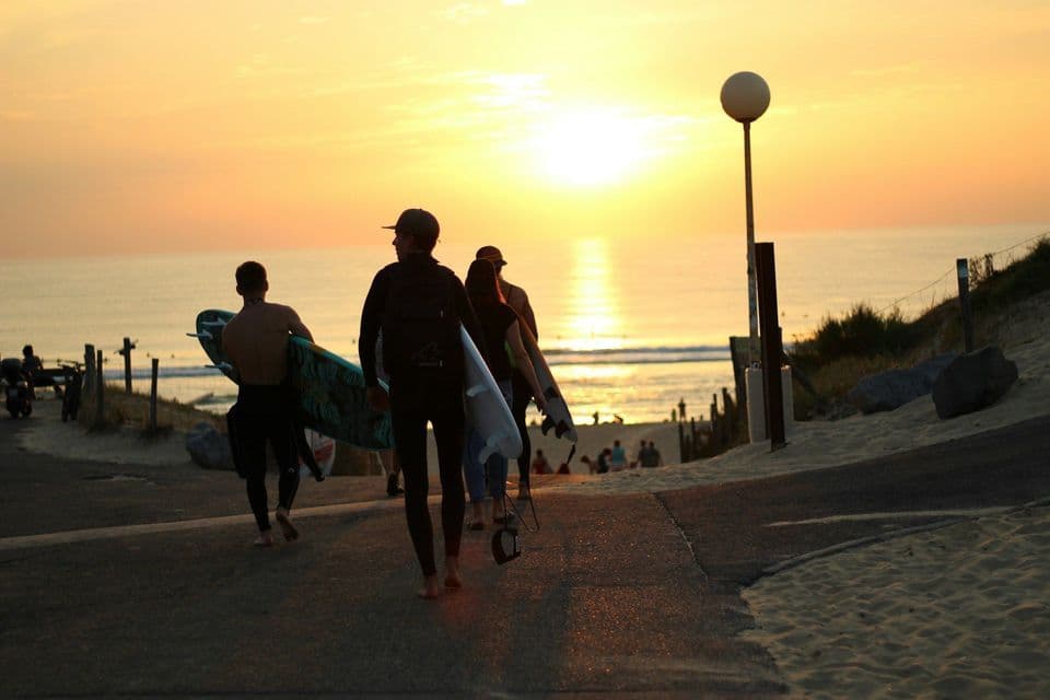 Un grupo de surfistas de WeRoad, con sus trajes de neopreno y tablas, camina por un sendero pavimentado hacia el océano al atardecer.