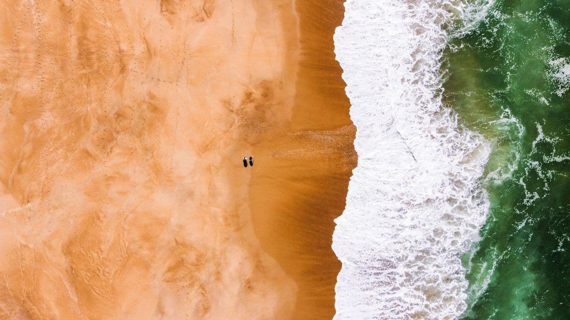 Una vista aérea cenital de dos personas de pie en una ancha playa de arena naranja junto al agitado mar verde.