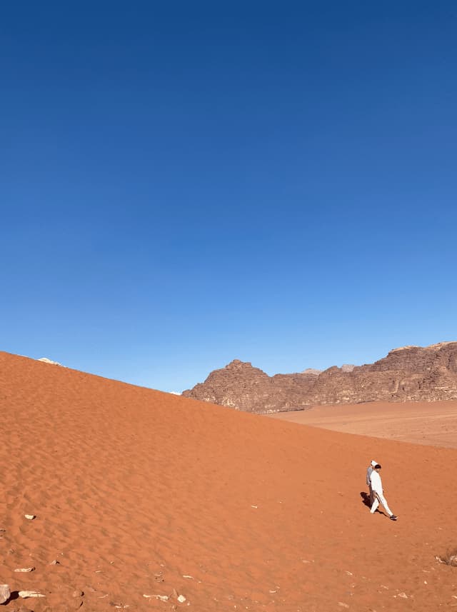 Una persona in abiti bianchi cammina lungo una vasta duna di sabbia rossa con montagne rocciose sullo sfondo sotto un cielo azzurro e limpido.