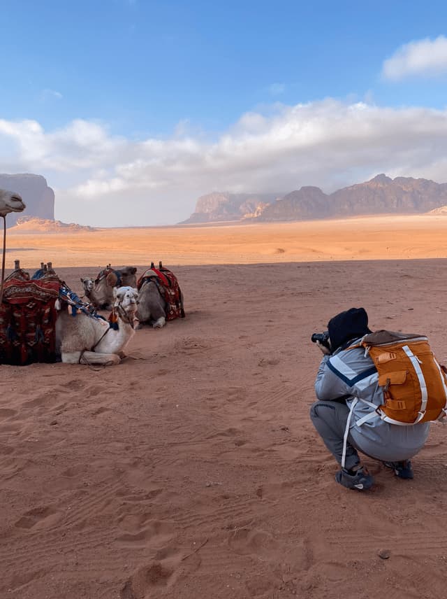 Una persona con uno zaino arancione si accovaccia sulla sabbia rossa del deserto per fotografare un gruppo di cammelli a riposo con le montagne sullo sfondo.