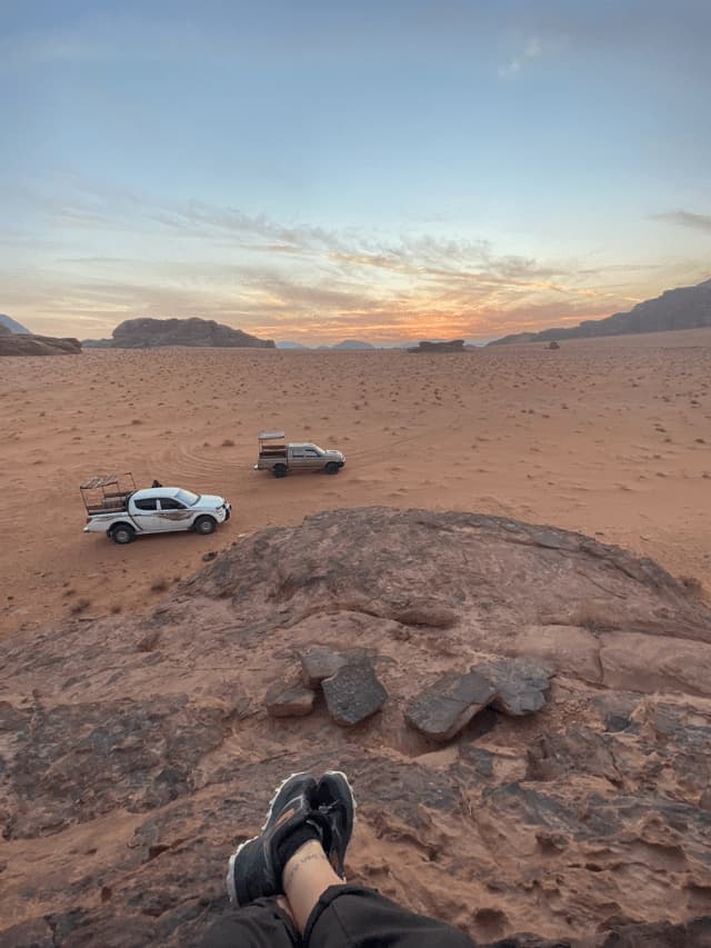 Una vista in prima persona da un affioramento roccioso mostra due camion da safari che attraversano un paesaggio desertico al tramonto.