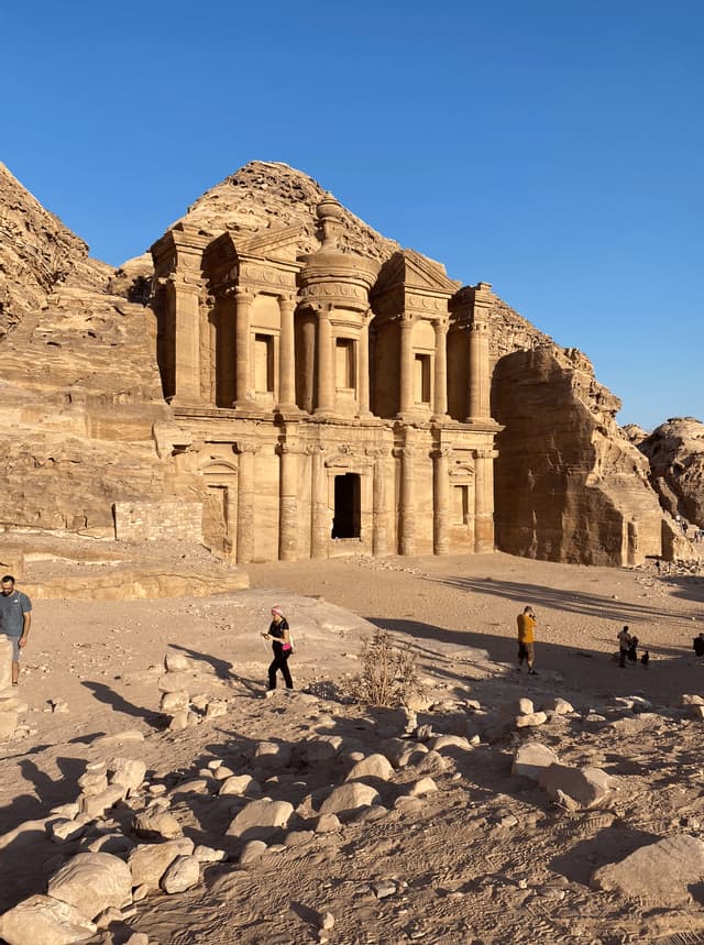 Un tempio monumentale scavato nella scogliera di arenaria, con persone in primo piano tra le rocce sotto un cielo azzurro e limpido.