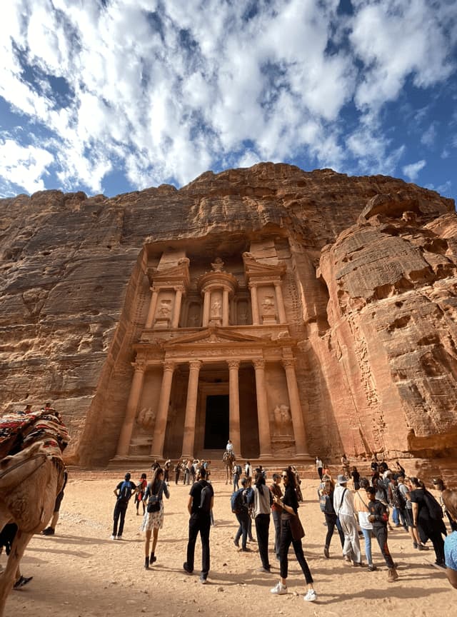 Un viaggio di gruppo WeRoad per ammirare il monumento di Al-Khazneh, una facciata scolpita in un'imponente scogliera di roccia rossa sotto un cielo parzialmente nuvoloso.