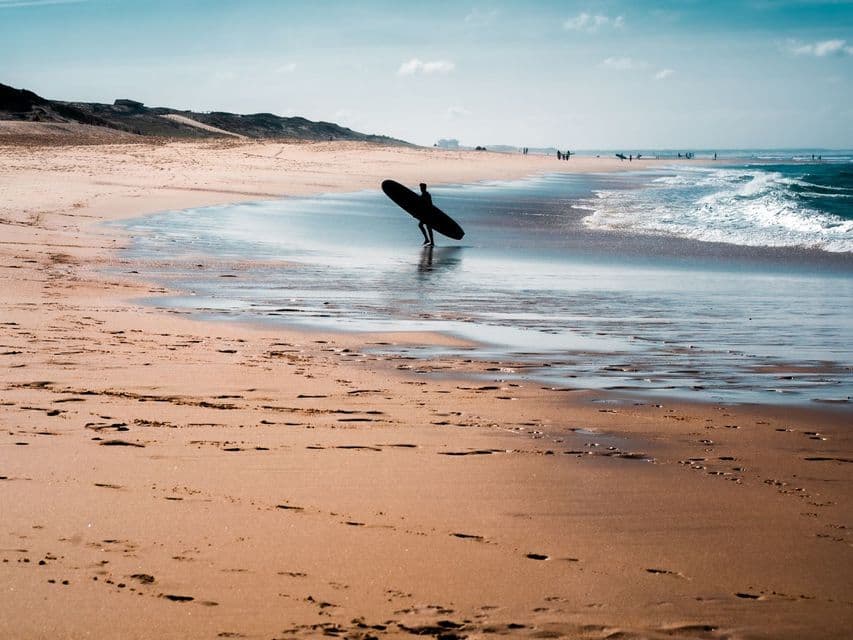 Un surfista en silueta con una tabla de surf camina hacia el agua en una playa ancha y arenosa