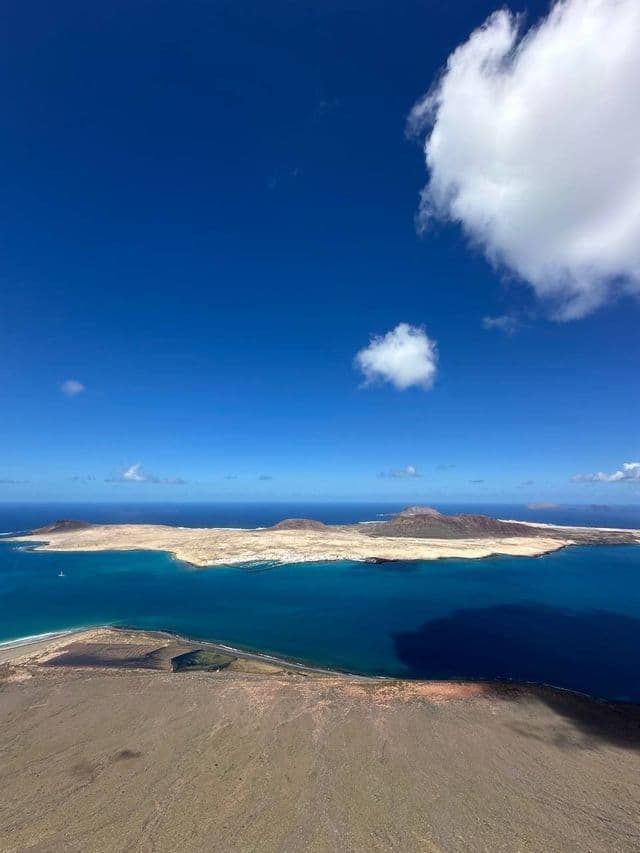 Una vista aerea di un'isola vulcanica sabbiosa circondata da acqua blu profonda sotto un cielo azzurro brillante con nuvole bianche sparse.