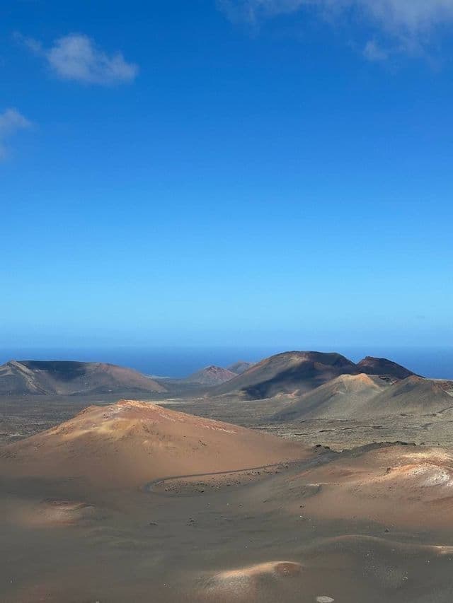 Una vista dall'alto di un paesaggio vulcanico con coni marrone-rossastro e terreno scuro, che conduce all'oceano sotto un cielo azzurro chiaro.