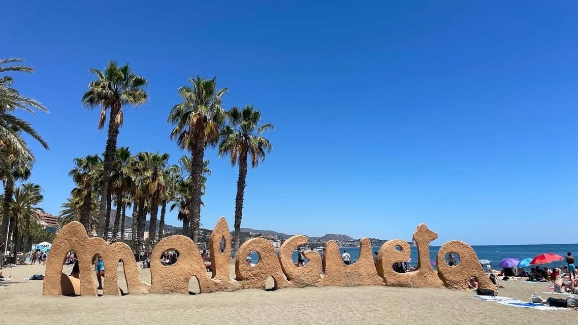 A sand-colored sculpture spelling 'Malagueta' stands on a sunny beach with palm trees and a clear blue sky.