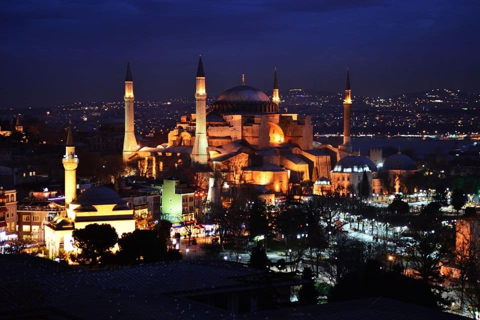 Una grande moschea con cupole e minareti, illuminata da una luce dorata contro lo skyline notturno di una città.