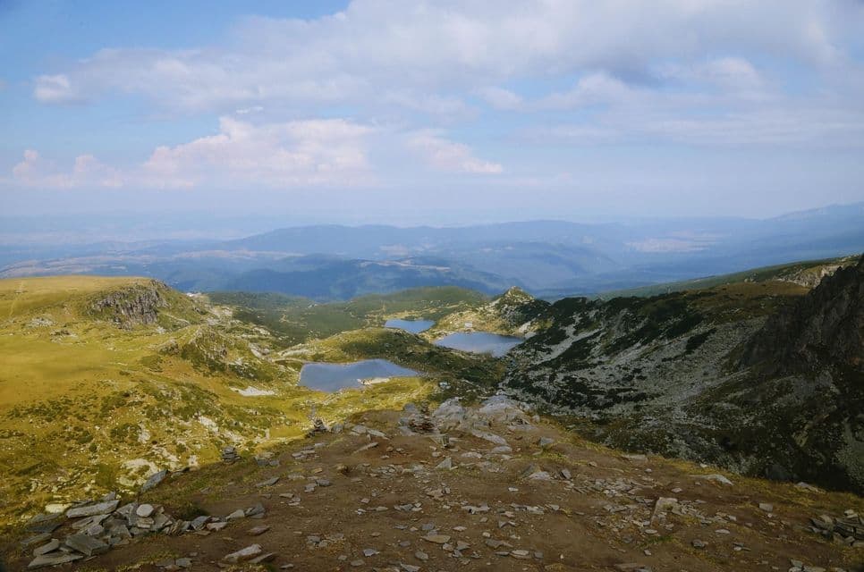 Vista da uno sperone roccioso che si affaccia su una verde valle montana con diversi laghi e un cielo blu nuvoloso.