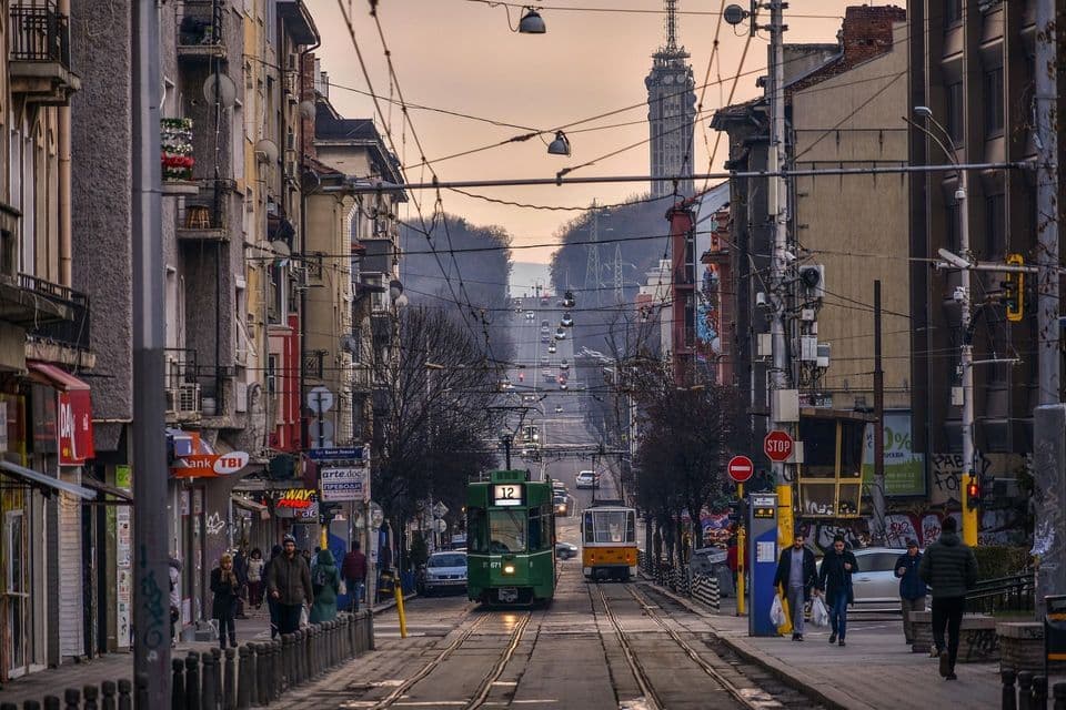 Un tram verde corre sui binari lungo una strada cittadina fiancheggiata da edifici, con un'alta torre di comunicazione visibile in lontananza.