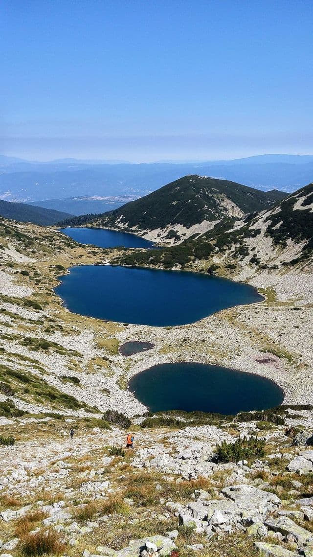Due partecipanti a un viaggio di gruppo WeRoad fanno trekking su un sentiero roccioso, con vista su diversi laghi alpini di un blu intenso, in una valle di montagna.