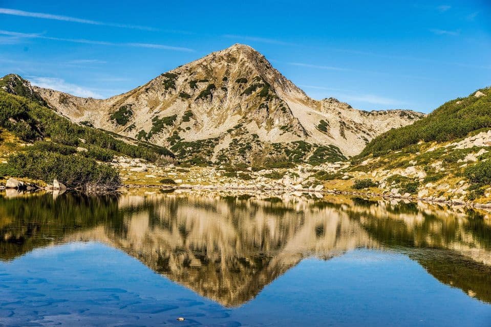 Una cima rocciosa di montagna si riflette nell'acqua calma e limpida di un lago in una giornata di sole.