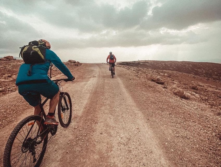 Due persone in mountain bike su una strada sterrata che si snoda attraverso un paesaggio roccioso e brullo sotto un cielo nuvoloso.