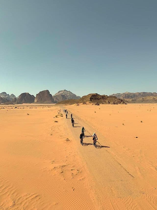 Un gruppo WeRoad in viaggio pedala in bicicletta in fila indiana lungo una strada sterrata attraverso un vasto deserto con montagne rocciose sullo sfondo.