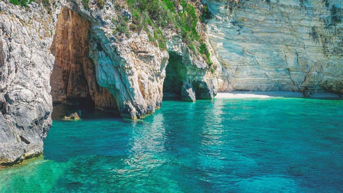 Acqua turchese si insinua in diverse grotte marine alla base di una grande scogliera rocciosa con una piccola spiaggia nascosta.