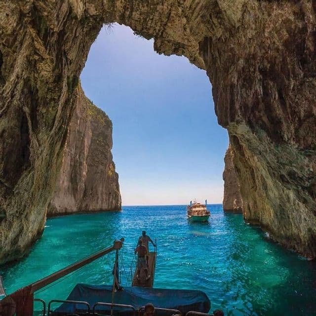 Una vista dall'interno di un'ampia grotta marina incornicia barche su acqua turchese vibrante circondate da ripide scogliere sotto un cielo soleggiato.