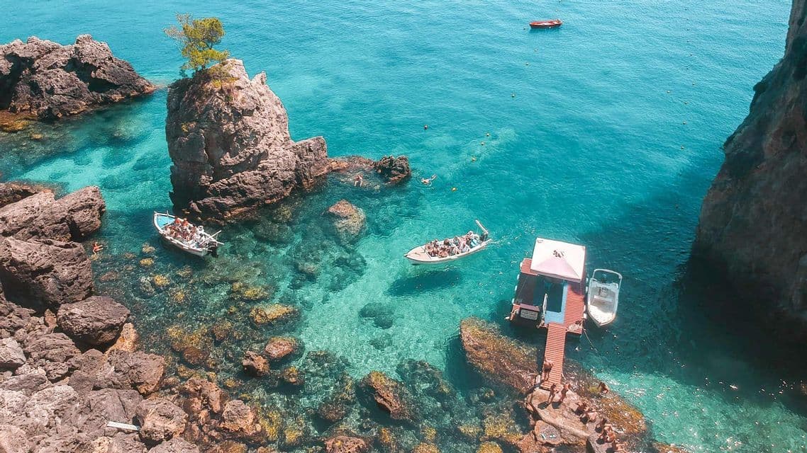 Veduta aerea di un viaggio di gruppo WeRoad in barca in una cala rocciosa con acqua cristallina turchese, un piccolo molo e nuotatori.