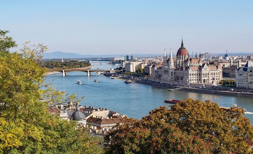 Une vue panoramique d'une ville avec un grand bâtiment orné sur la rive d'une rivière, avec des bateaux sur l'eau sous un ciel bleu clair.