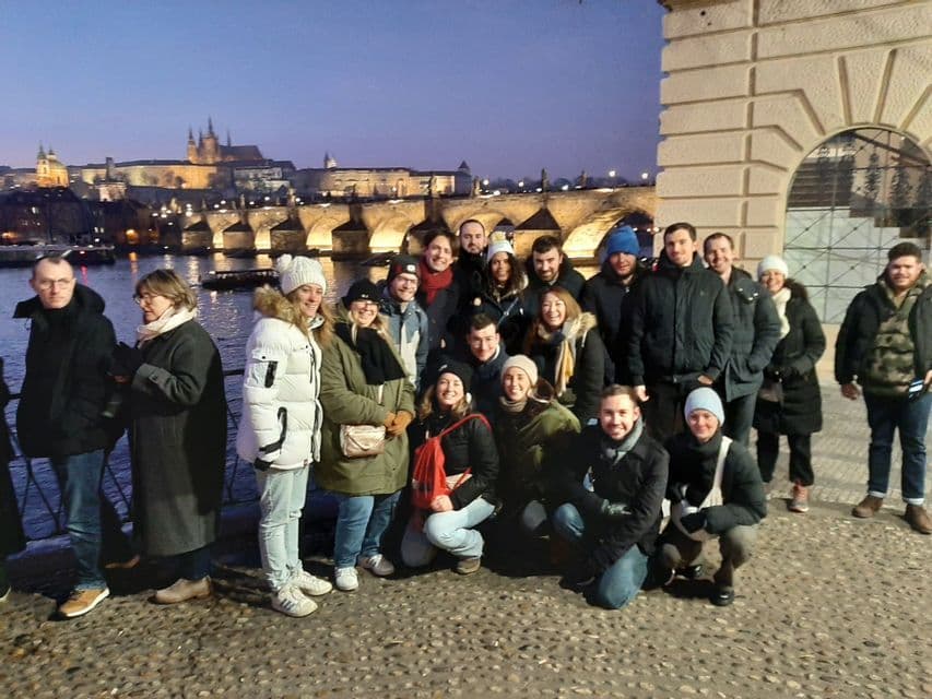 Un groupe WeRoad en habits d'hiver pose sur un chemin pavé au crépuscule, avec un pont illuminé au-dessus d'une rivière en arrière-plan.