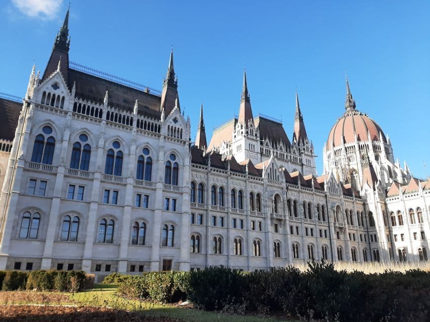 La façade ornée d'un grand bâtiment avec des flèches gothiques et un dôme central, vue depuis une pelouse sous un ciel bleu clair.