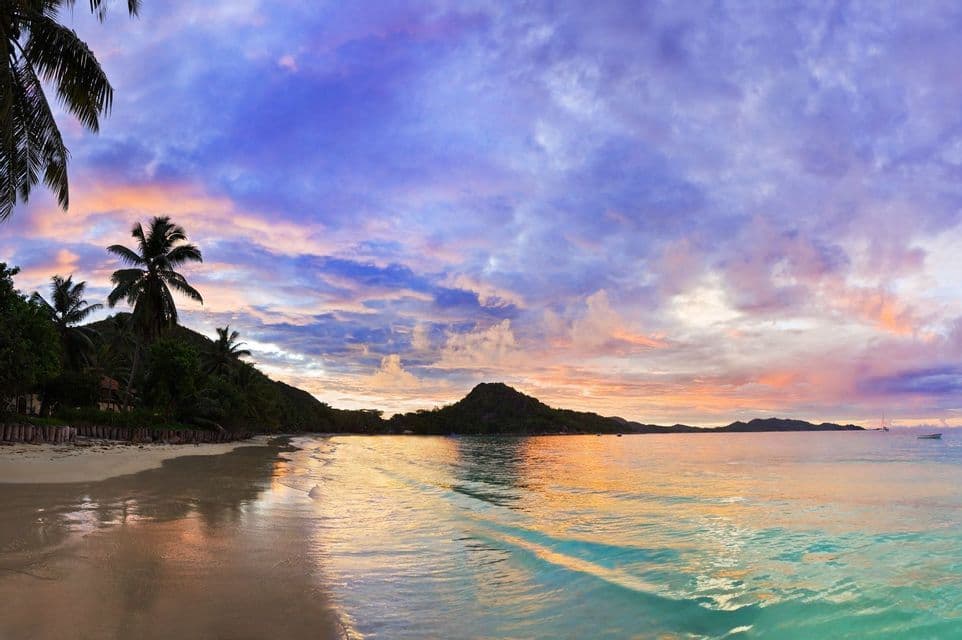 Ein tropischer Panoramastrand bei Sonnenuntergang, mit lila und orangefarbenen Wolken, die sich auf dem ruhigen Ozean spiegeln.