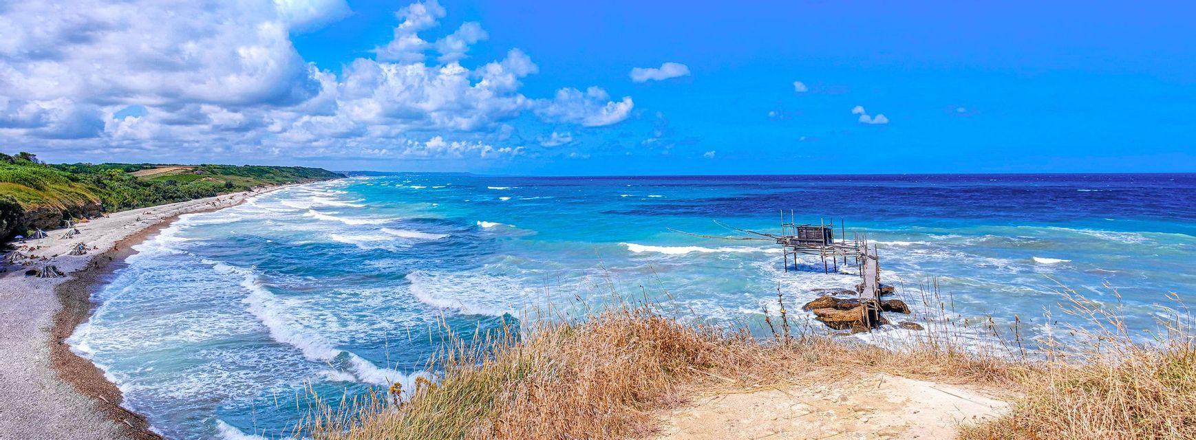 Una vista panoramica di una costa con un pontile da pesca in legno nel mare turchese, accanto a una lunga spiaggia e colline verdi.