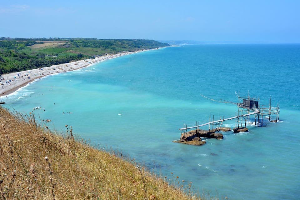 Vista dall'alto di una costa con una lunga spiaggia, acqua turchese e un tradizionale molo da pesca in legno su palafitte.