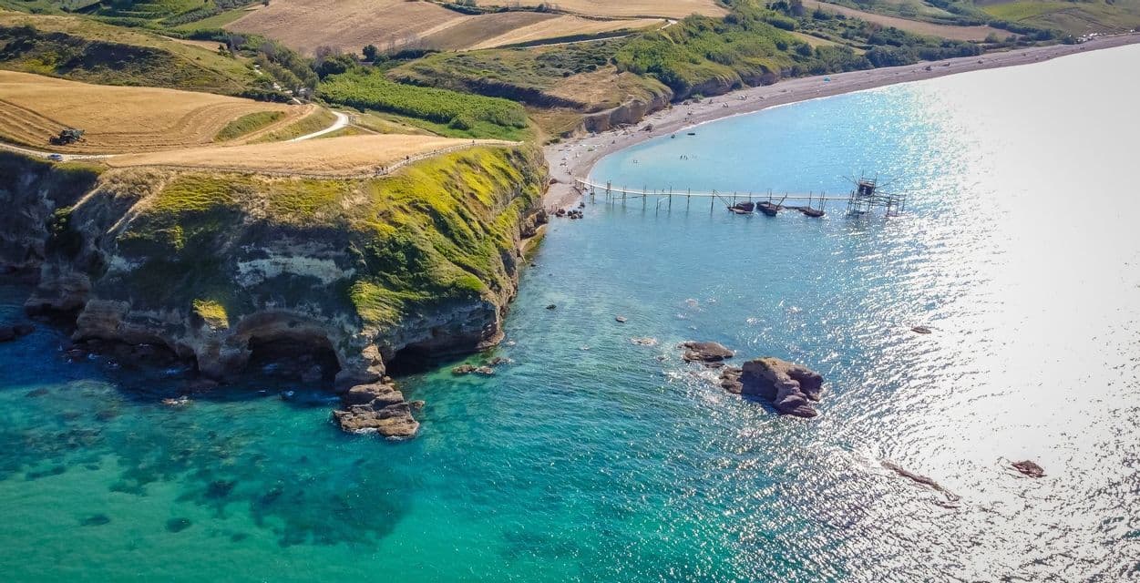Veduta aerea di un pontile in legno che si protende da una costa rocciosa e rigogliosa verso un mare turchese, con dolci colline alle spalle.