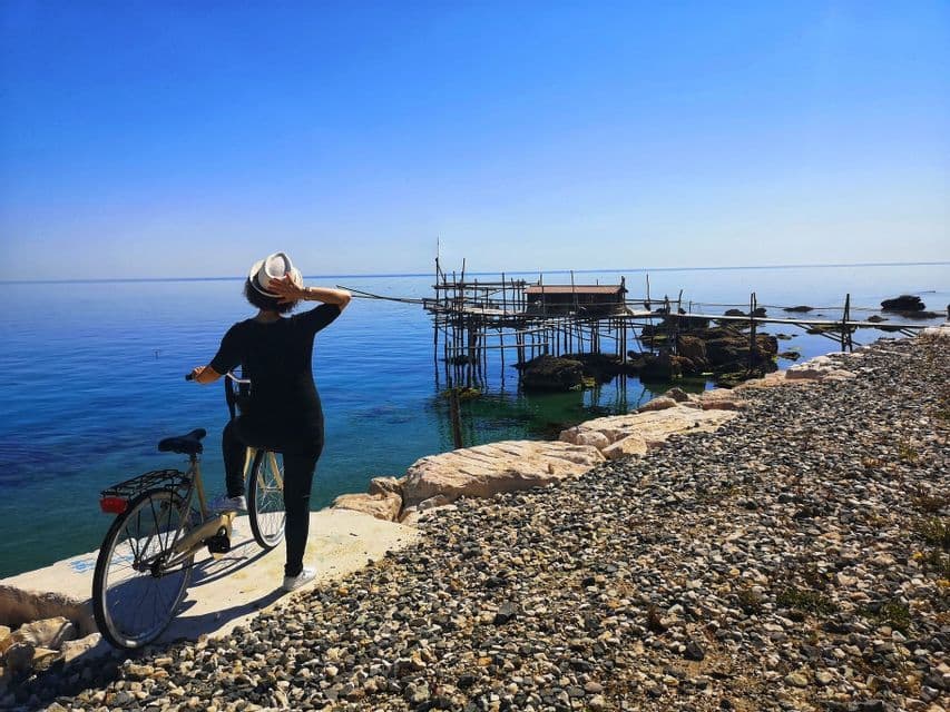 Una persona con un cappello bianco si trova con una bicicletta su una costa rocciosa, osservando un molo di legno per la pesca sul mare calmo e blu.