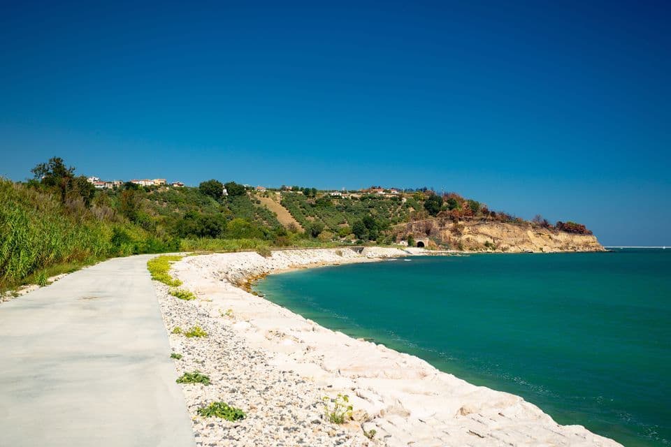 Un sentiero costiero pavimentato corre lungo una riva rocciosa accanto a un'acqua turchese, con una lussureggiante collina verde sotto un cielo azzurro.