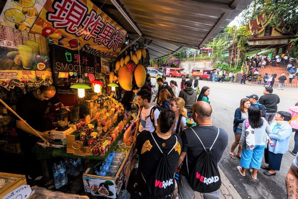 A WeRoad group trip gathers at a bustling outdoor fruit smoothie stall while a vendor prepares drinks.