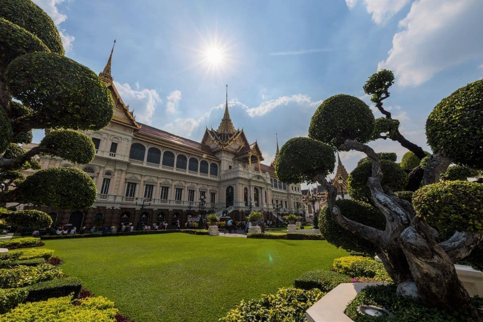 Un palais orné au toit doré se dresse derrière une vaste pelouse impeccablement entretenue, encadré par des arbres taillés en topiaire sous un soleil éclatant.