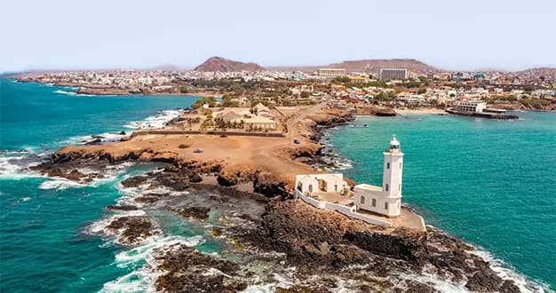 Eine Luftaufnahme eines weißen Leuchtturms auf einer felsigen Halbinsel, die in türkisfarbenes Wasser ragt, mit einer Küstenstadt im Hintergrund.