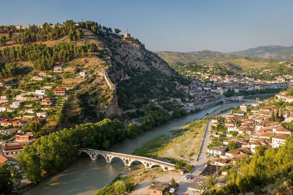Un puente de piedra arqueado cruzando un río en un pueblo histórico enclavado en un valle con una colina fortificada.