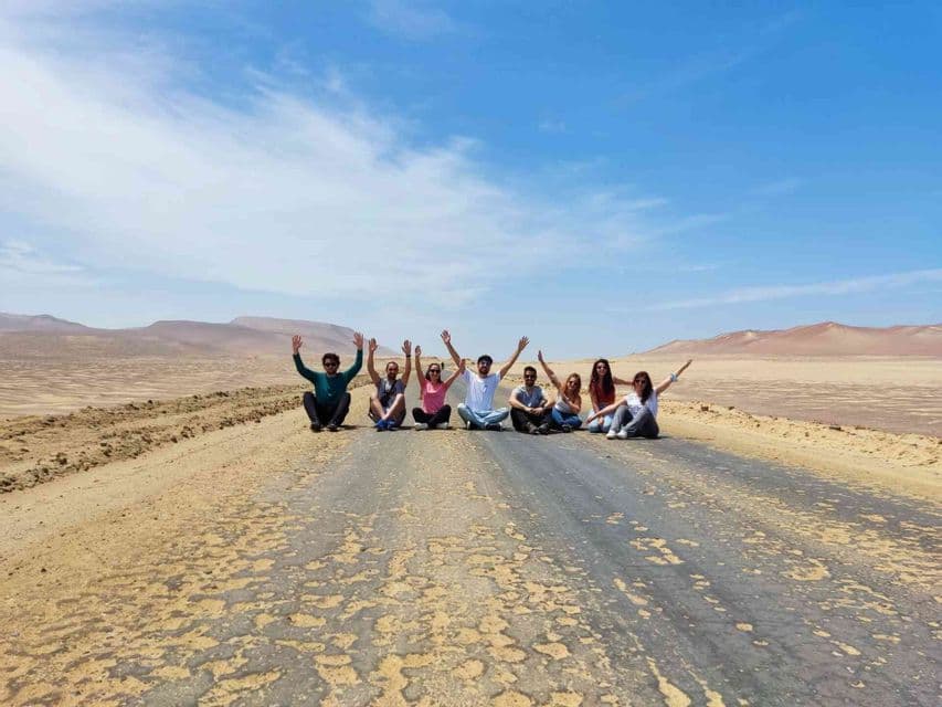 Un grupo de WeRoad en viaje sentados en una carretera erosionada en un vasto desierto, con los brazos alzados hacia un cielo azul.